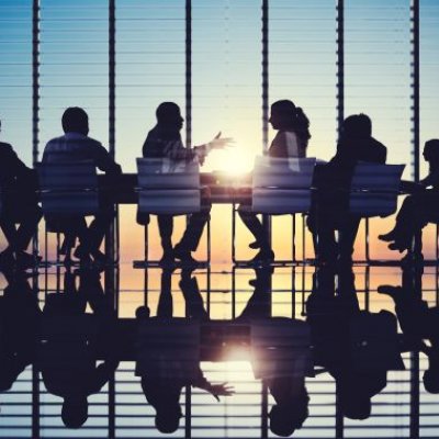 Men and women sitting around a board room table with sun shining through glass wall. Adobe